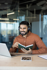 Young indian man wearing headphones and holding a notebook while having an online lesson or working remotely on a laptop in a contemporary office environment