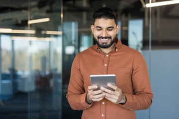Young man smiling. Wearing an orange shirt. Standing in a modern office space while engaging with a digital tablet. Representing technology. Communication. And connectivity for business professionals