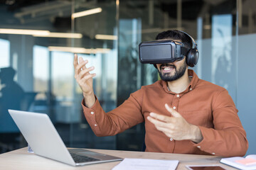 Young man wearing vr headset and headphones, interacting with virtual content while sitting at a desk with a laptop, embracing metaverse technology in a corporate environment