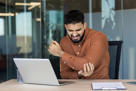Young indian businessman clutching his elbow in pain while working on a laptop at his desk, suggesting repetitive strain, tendonitis or chronic discomfort from sedentary office work