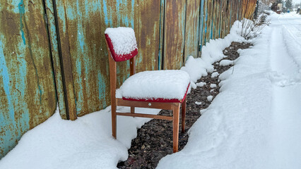 Old red upholstered wooden chair heavily covered in fresh snow, abandoned outdoors next to a rusty wall and a blue fence during a cold winter day.