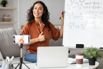Female teacher recommending online school, showing thumb up gesture sign, giving virtual lesson, explaining math. Positive young tutor satisfied with educational services, recording video on cellphone