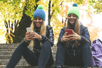 Two girls with wool cap sitting in the park using smartphone, beautiful autumn afternoon. Teen using mobile phone. Sisters chat with friends and classmates. Young women enjoying free time in nature