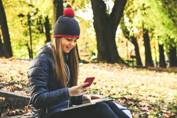 Girl with wool cap sitting in the park using smartphone, beautiful autumn afternoon. Teen using...