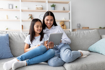 Young asian mother and daughter sitting on sofa and using digital tablet, watching videos or surfing internet, copy space. Positive family using mobile app on pad together © Prostock-studio