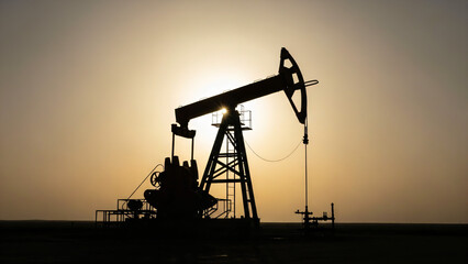 A silhouette of an oil pump jack is centered against a bright, hazy sky, likely at sunrise or sunset. Large mechanical device used for extracting crude oil from an oil well dominating the frame.
