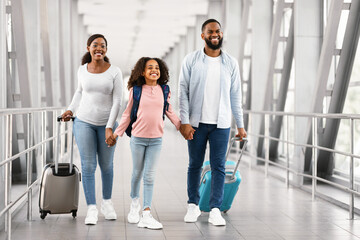 Fototapeta premium Family Travel Concept. Portrait of excited black dad, mum and girl walking at airport terminal corridor with suitcases waiting for the aircraft arrival, three people holding hands, free copy space