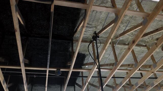 Construction workers spraying black paint on ceiling joists with foam insulation underneath.