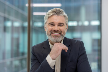 Experienced senior businessman with grey hair and beard smiling confidently at camera in a modern corporate office, conveying leadership, professionalism and approachability