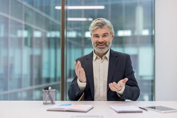 Mature businessman with a grey beard and suit smiling while sitting at an office desk, actively gesturing with hands during a virtual meeting or online presentation © Liubomir