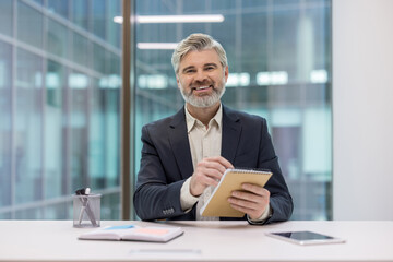 Confident senior businessman with gray hair and beard sitting at a modern office desk, holding a notebook and pen, smiling at the viewer while actively engaged in planning or brainstorming
