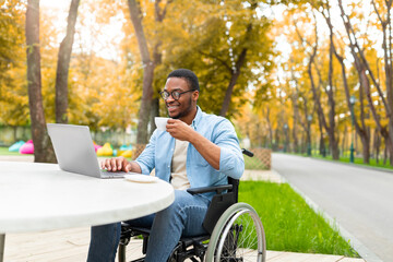 Remote job for impaired people. Cheerful black guy in wheelchair working online, using laptop at outdoor cafe in autumn. Disabled young Afro man studying, attending webinar, drinking coffee at park