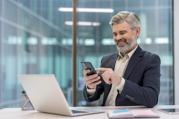 Senior businessman smiling while texting on a mobile phone, sitting at his desk with a laptop and planner in a contemporary office building, representing modern communication and business technology