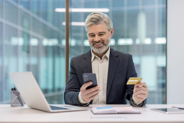 Smiling mature businessman at office desk holding credit card and smartphone while completing an online payment or e-commerce purchase, confident and relaxed during internet banking