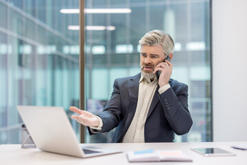 Mature businessman experiencing stress during a frustrating phone call while looking at his laptop, dealing with problems in a modern office environment