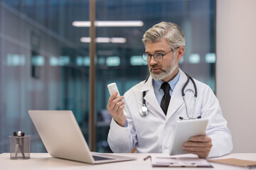 Middle-aged male doctor in lab coat and stethoscope holding a medicine bottle, looking at it intently while also accessing patient information on a digital tablet at his office desk with a laptop