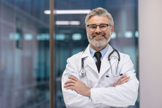 Experienced physician with gray hair and beard standing with crossed arms in a hospital corridor, representing medical care, professionalism, and trust in healthcare