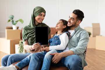 Great Website. Smiling muslim dad, mom and daughter sitting on the sofa chair in living room among carton boxes after relocation, woman showing digital tablet with black empty screen for mock up