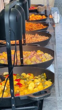 outdoor paella pans at market rows of giant skillets filled with saffron rice, colorful peppers, shrimp and chorizo, steam rising, ladle stirring, Vertical shot taken on mobile device.