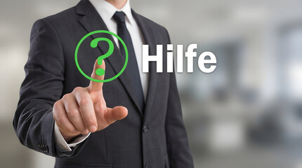 Businessman in dark suit points to virtual help icon with green question mark and German text Hilfe on bright blurred office background signifying gui