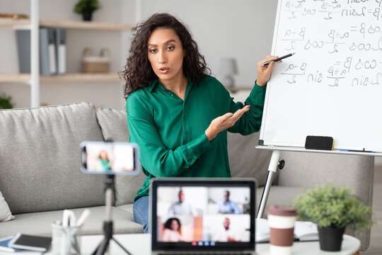 Online Studying, Distance Learning, Virtual Class. Serious focused female teacher shooting lesson on smart phone camera, sitting at desk near flipchart, talking to pc screen with diverse students - Powered by Adobe