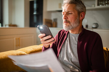 Mature man concerned using smartphone flashlight to read bills at home