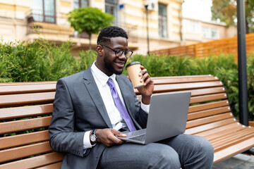 Corporate african american executive in formal wear sitting on bench near office center with laptop and takeout coffee during lunch break. Happy black IT manager getting ready for business meeting