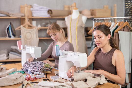 Two female tailors are sitting at a table in the workshop and sewing clothes on machines. Employees of the sewing industry make wear according to individual measurements