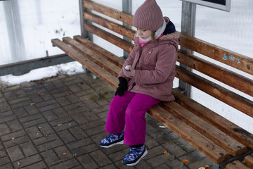 little girl sitting on bench using tablet in covered shelter, bundled in pink jacket and mittens,...