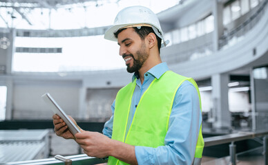 A worker wearing a safety vest and hard hat stands in a large industrial setting. He looks at a tablet, focusing on the screen as he engages with the device.