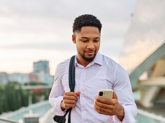 Portrait of a young businessman man using a smartphone mobile phone walking down the street, surrounded by moder corporate office buildings and modern city architecture