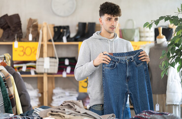 Regular customer guy client pre-examine fabric and style of product, is busy choosing jeans trousers. Client enjoys shopping for clothes at store browsing through colorful on display racks