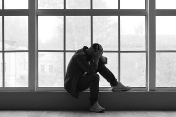 Fototapeta premium Depressed young man sitting near window at home. International Stress Awareness Week