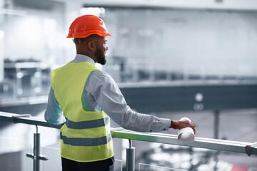 A worker in a safety vest and helmet stands at a railing, looking out over a construction site. He holds a rolled-up plan in one hand and seems to be focused on the work below.