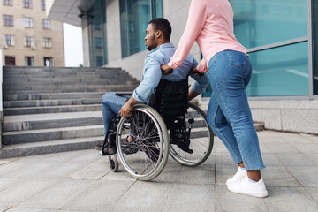 Obraz premium Problems with accessible environment for people with physical disability. Young black woman with millennial man in wheelchair having no possibility to enter building without ramp, outdoors