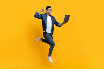 Emotional arab young man jumping up, using new modern laptop, raising fist up and smiling over yellow studio background, trading online, copy space for text or advertisement, panorama