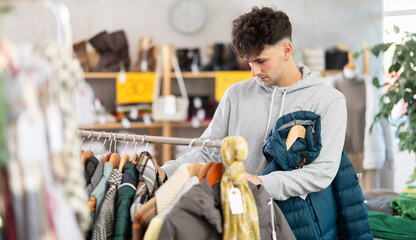 Young man in twenties updating wardrobe, carefully choosing winter puffer jacket in local clothing store, checking insulation and material before buying