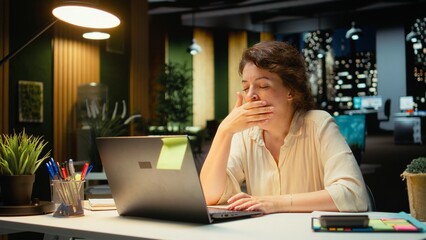 Restless sleepy businesswoman yawning in the office struggling to stay awake, dealing with sleep deprivation and work tension due to overload at night. Pushing limits with discomfort. Camera A.