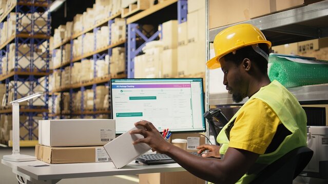 Black depot clerk reviewing inventory data on tracking software for distribution system, busy logistics hub stock room with organized racks. Online retail fulfillment and supply chain. Camera A.
