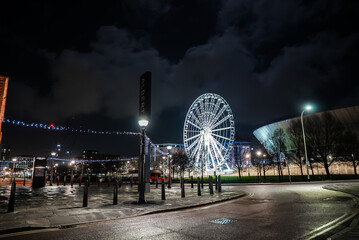 Obraz premium Nighttime Liverpool waterfront with Liverpool Wheel spinning by ACC Liverpool and M and S Bank Arena. Festive lights line Albert Dock promenade under a dark clouded sky.