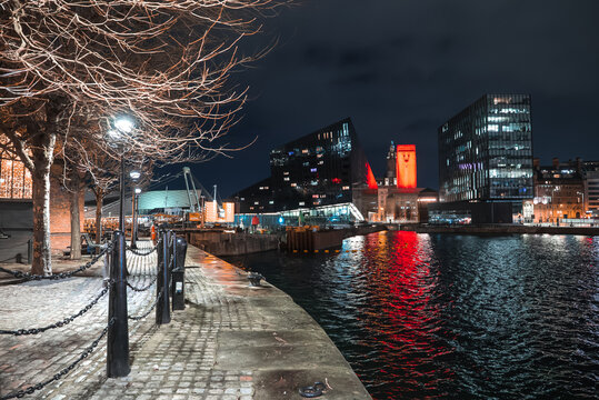 Nighttime Liverpool waterfront shows Mersey promenade, cobblestones, chain bollards, bare winter trees, Mann Island, Museum of Liverpool, and Port clock tower glowing red.