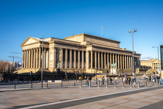 St George's Hall in Liverpool, England, shows a long colonnade, equestrian statues, and a Union Jack. People in winter coats cross the plaza under a crisp blue sky.