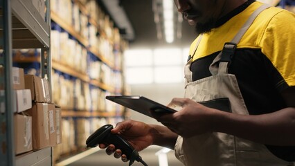 Black male worker using scanner checking airway bill numbers in inventory, solving logistics operations in industrial warehouse. Worker verifying shipping labels near shelving units. Camera B.