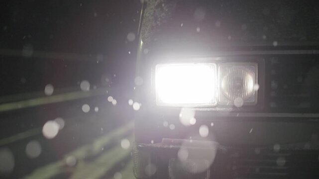 A close-up of a vintage vehicle s rectangular headlight glowing intensely in the dark. Snowflakes fall in the foreground, creating soft, blurry white bokeh circles against the moody night atmosphere.