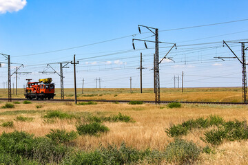 Railway maintenance train working on tracks in open steppe landscape, surrounded by power line poles. Symbol of repair works, railway infrastructure, rail transportation, and heavy industry.