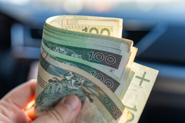 Hand holding folded stack of Polish zloty 100 banknotes with blurred car interior dashboard in background. Concept of driver earnings, cash savings, fuel budget and automotive financial transactions. 