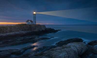 Lighthouse on rocky coastline at dusk with beam of light.