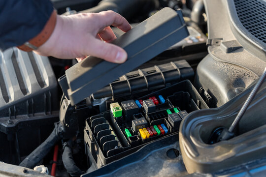 Male hand lifting plastic cover of automotive fuse box revealing colorful blade fuses and relays in car engine compartment. Concept of electrical system inspection, vehicle diagnostics and DIY repair.