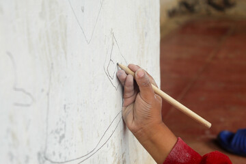 The hand of a little boy is learning to draw on a white wall using a simple wooden pencil.
