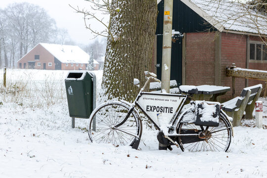 Announcement notice for exposition on an old fashioned bicycle stationed against a tree covered in a thick layer of snow after snowstorm blizzard in Dutch countryside with bench in the background